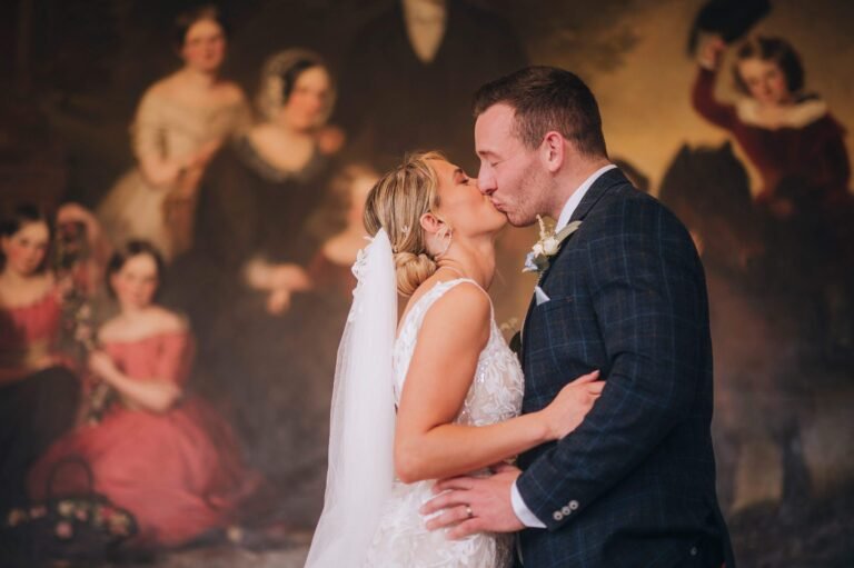 A bride and groom share a kiss in front of a historical painting with figures in 19th-century attire, captured beautifully by a Norwood Park wedding photographer. The bride stuns in her lace gown and veil, while the groom looks dashing in his dark suit with a boutonniere.