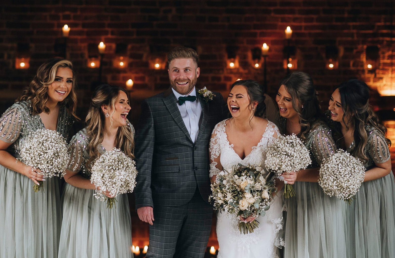 A groom and bride stand smiling with four bridesmaids holding bouquets of babys breath. The women wear matching dresses, and candles glow in the background against a dark brick wall, creating a warm atmosphere captured perfectly by their Nottinghamshire wedding photographer.