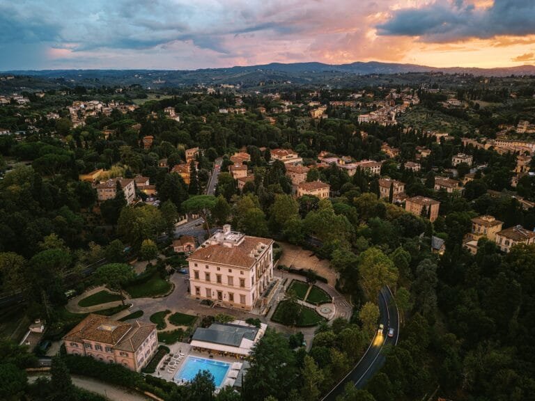 Aerial view of a sprawling landscape at sunset, showcasing Villa Cora surrounded by lush greenery and numerous trees. Several smaller buildings and a winding road with a car are visible, set against distant hills under a colorful sky—perfect for Florence destination wedding photography.