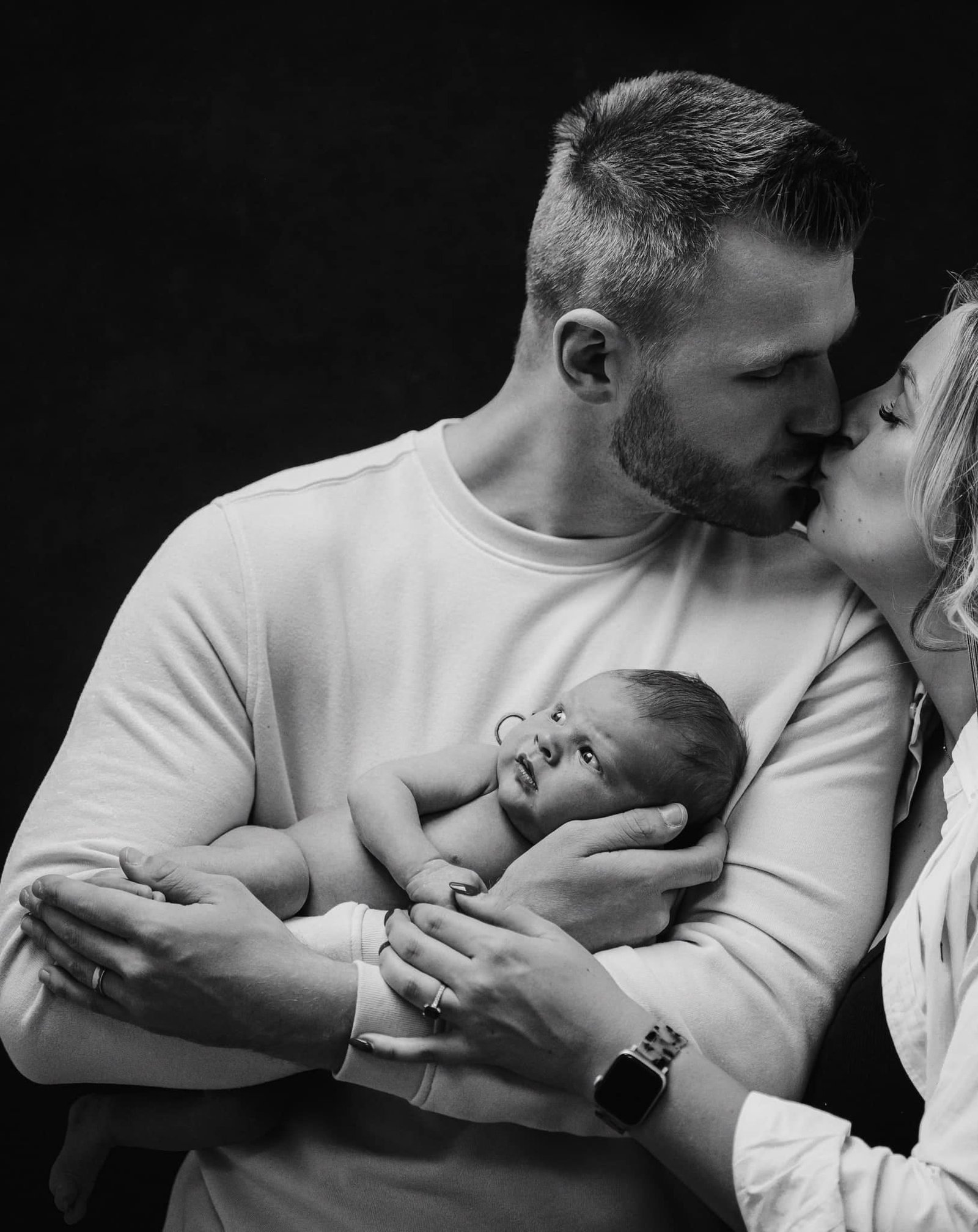 A black and white image captures a couple tenderly enveloping their newborn. The man, embodying the Yorkshire charm, kisses the woman’s forehead as he cradles their baby. Her gentle touch on the babys arm meets its calm upward gaze in this timeless photograph by a renowned newborn photographer.