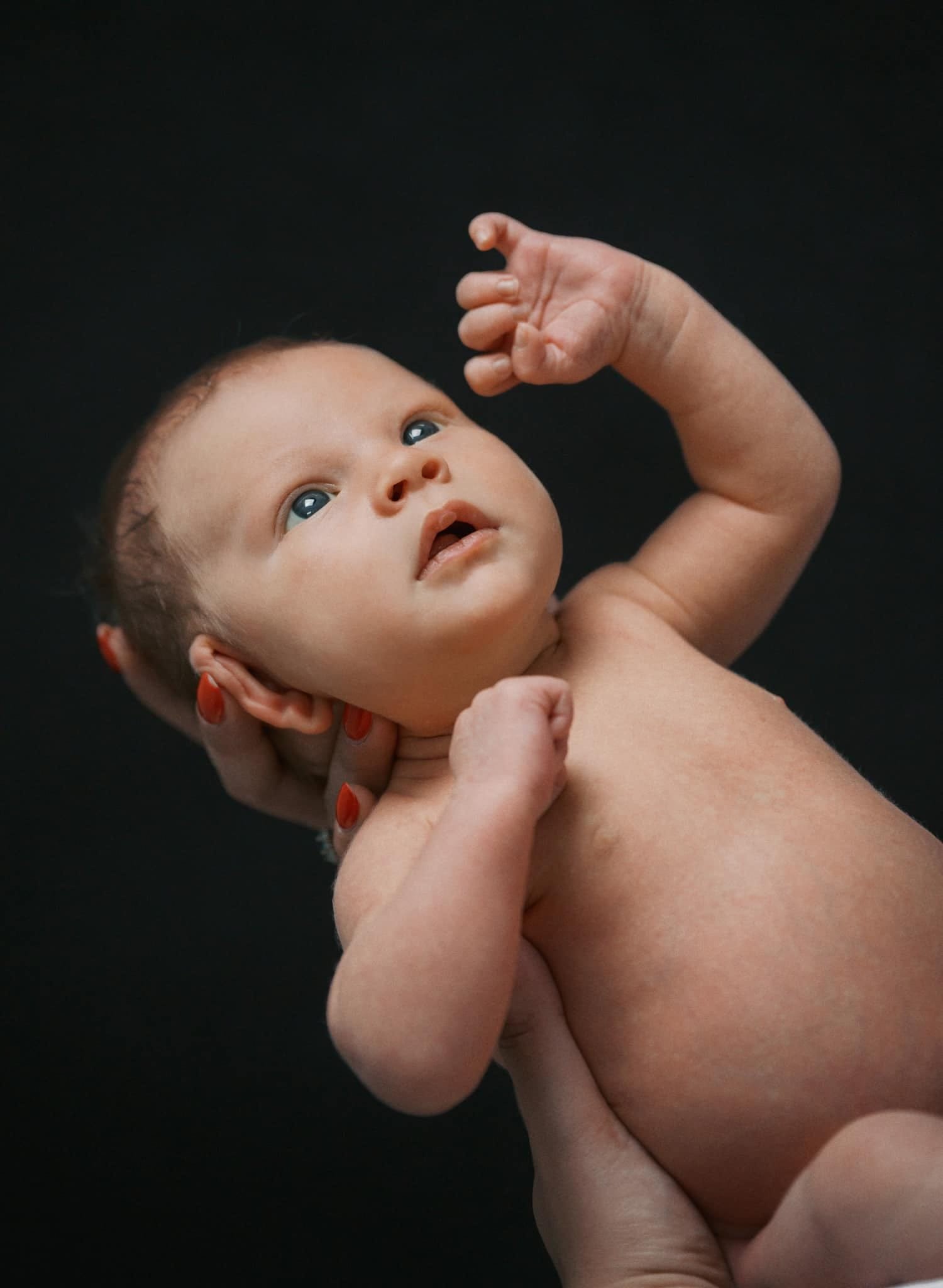 A newborn baby with light skin is gently cradled in adult hands by a Yorkshire newborn baby photographer. The babys curious expression and tiny, raised arms are beautifully captured against a dark background, highlighting its delicate features.