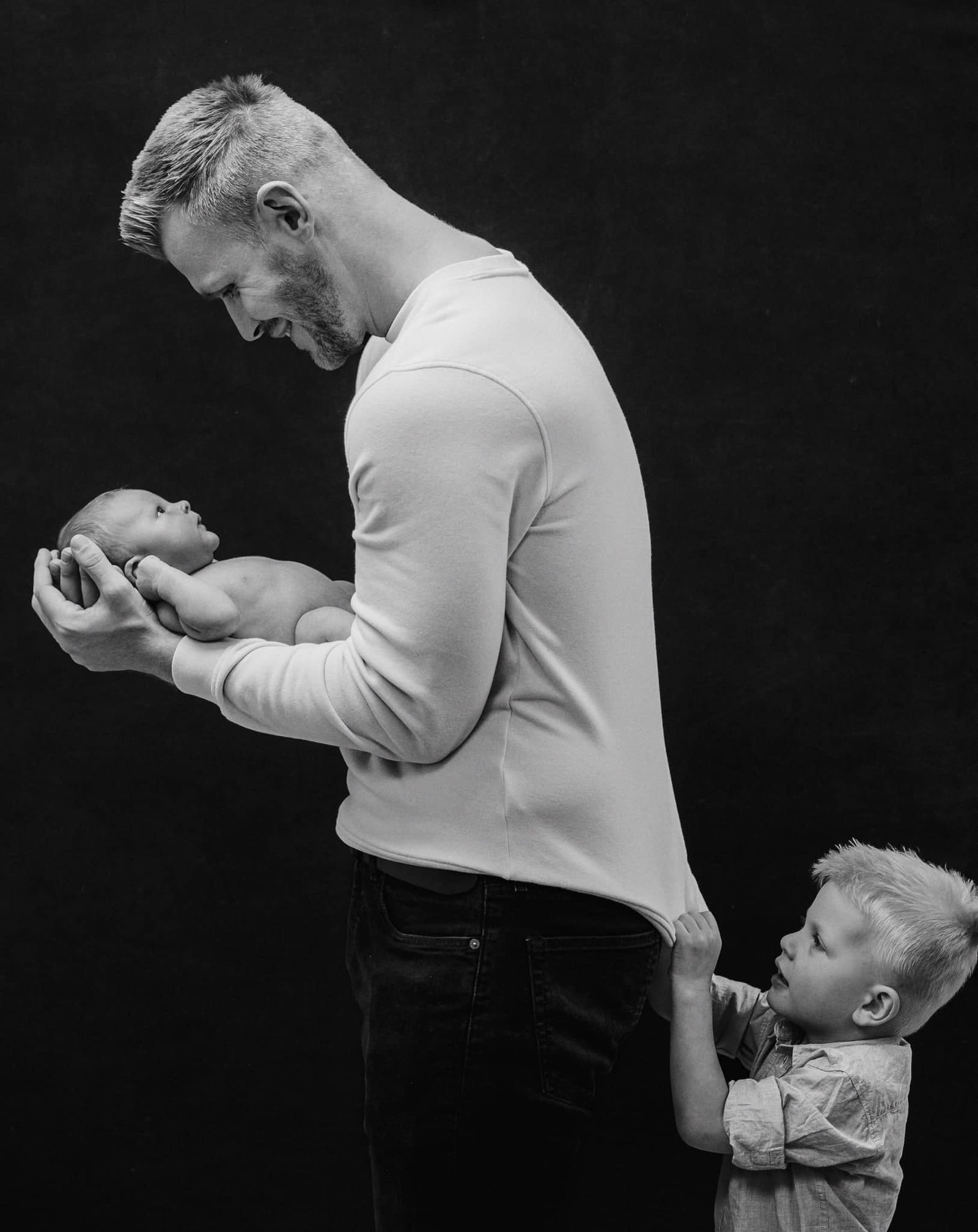 A black-and-white photo captures a man holding a baby in his arms, smiling gently—a tender family moment beautifully framed by the keen eye of a Yorkshire newborn baby photographer. A young child tugs on the mans shirt beside him, set against a plain, dark background.