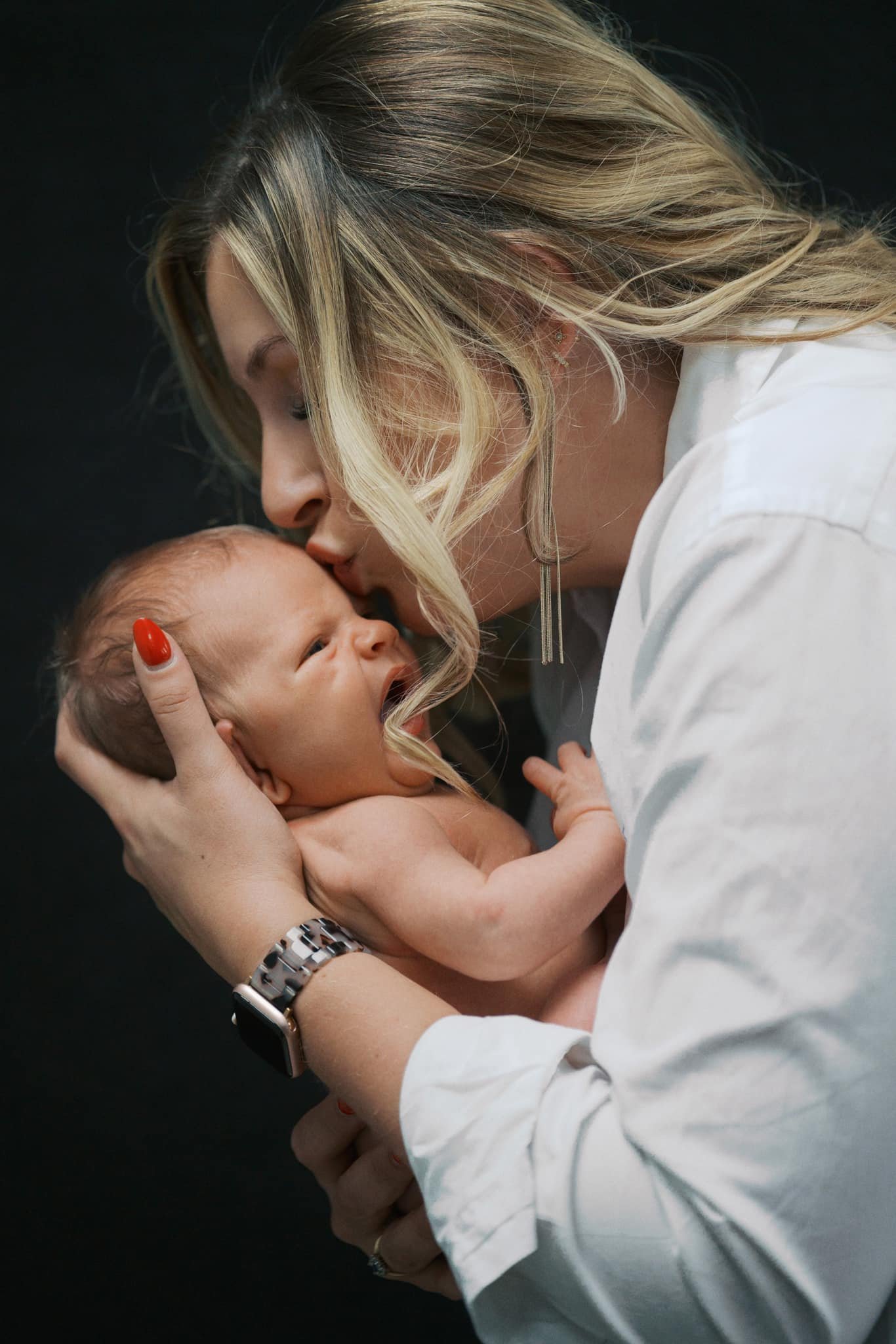 A Yorkshire newborn baby photographer captures a tender moment as a woman with long blonde hair gently kisses a newborn on the head, holding them closely. She wears a white shirt and watch, set against a dark background that draws all attention to their shared affection.