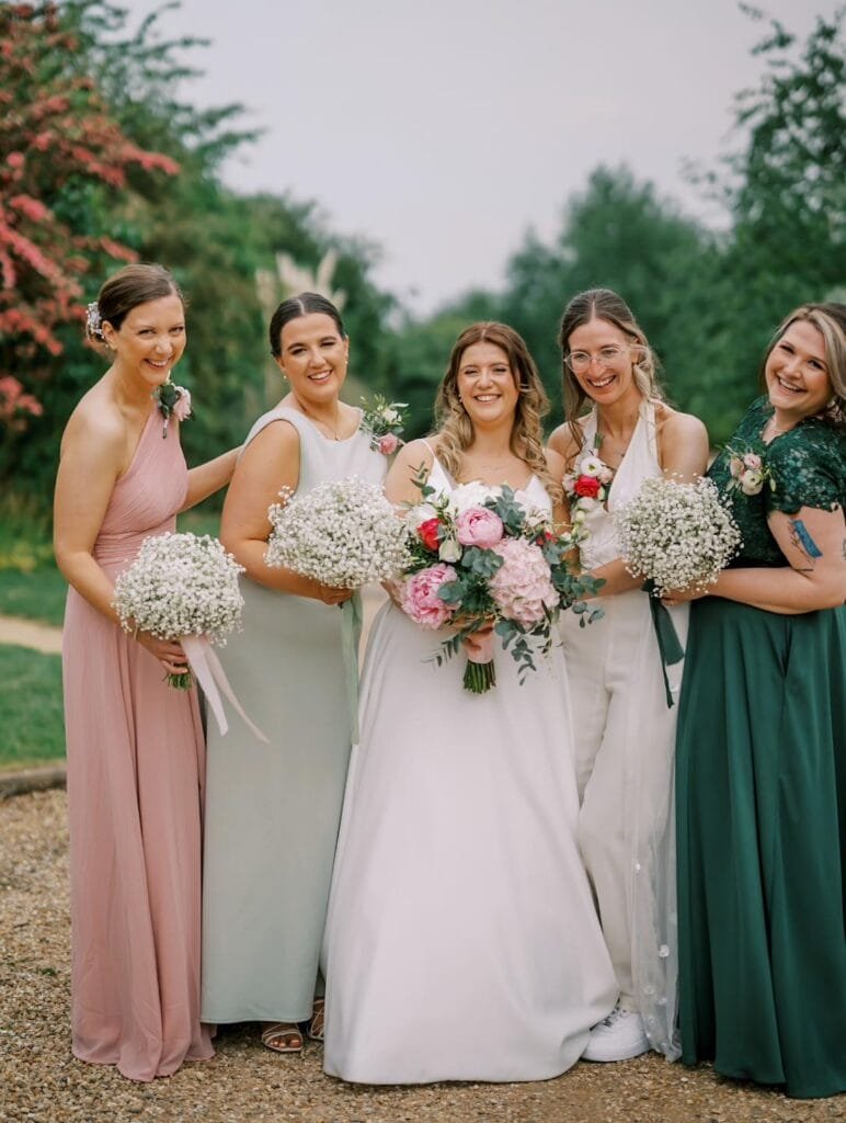 A bride in a white gown holds a pink and white bouquet, surrounded by four bridesmaids in pastel dresses. They smile outdoors on a picturesque path, with trees and flowers adding charm to the scene—perfect for a Carriage Hall wedding photo capturing the magic of this Nottinghamshire wedding venue.