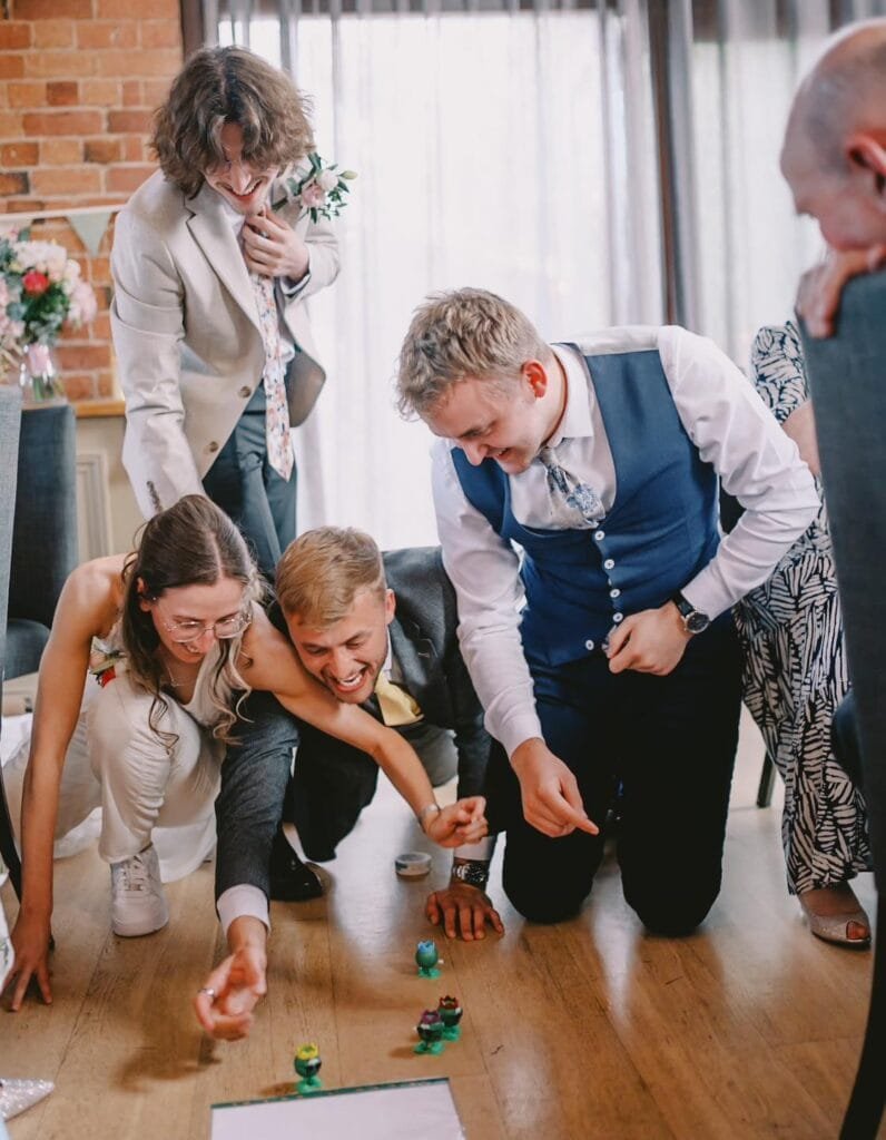 A group of people kneel on the floor, enthusiastically playing a game with plastic toy figurines amidst the vibrant atmosphere of a Nottinghamshire wedding venue. They are laughing and engaged, surrounded by bright lighting and a rustic brick wall backdrop.