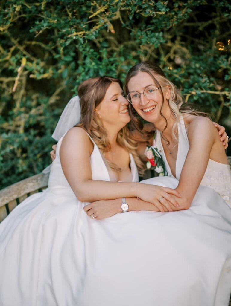 Two brides in white dresses sit close together on a bench, surrounded by greenery at a picturesque Nottinghamshire wedding venue. One smiles at the other, who looks at the camera with a soft smile and glasses. They hold hands, one adorned with a flower corsage—its a perfect carriage hall wedding photo.