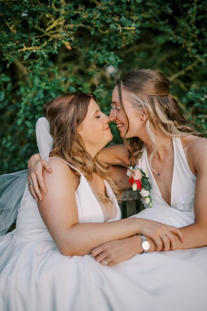 Two brides in wedding dresses share a joyful moment, sitting close with arms around each other. Both have long hair and glasses, smiling warmly. The outdoor setting features lush greenery, perfectly captured by a Carriage Hall wedding photographer at the scenic Nottinghamshire wedding venue.