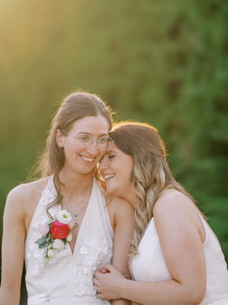 Two people in white dresses share a joyful moment outdoors at a Nottinghamshire wedding venue. One wears glasses and holds a bouquet with a red rose, while the other leans in, smiling. Sunlight softly filters through, and a lush green background frames this carriage hall wedding photo.