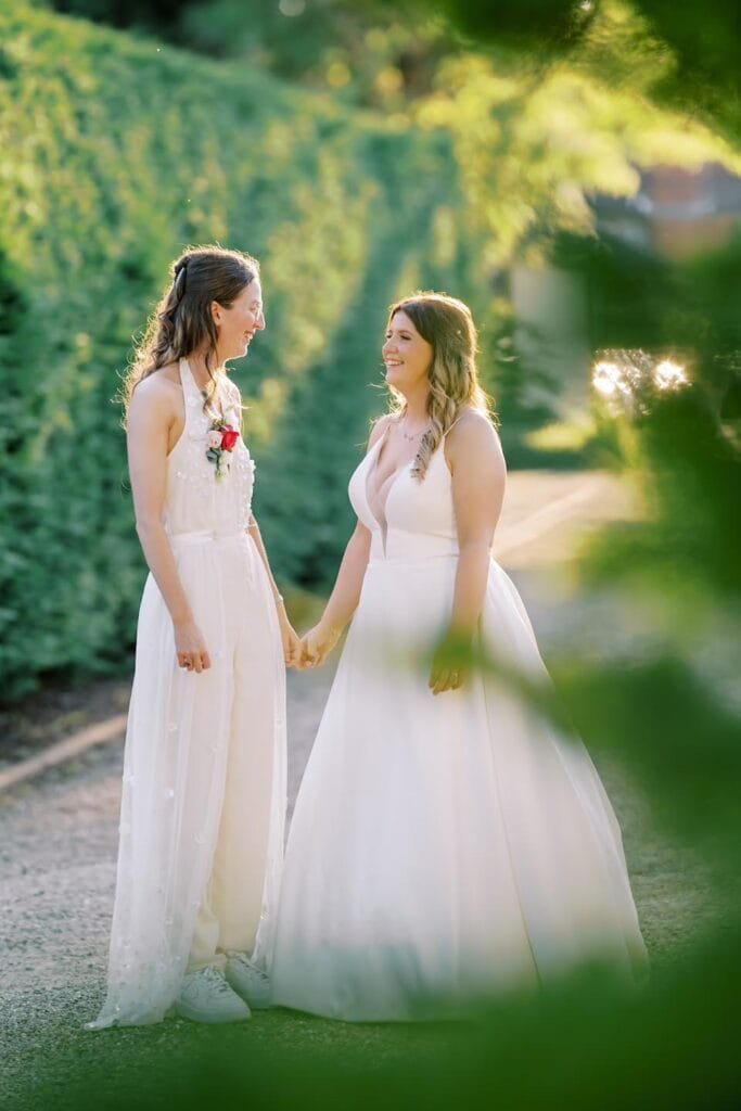 In the serene setting of a Nottinghamshire wedding venue, two women in wedding dresses stand hand in hand, smiling at each other amid lush greenery. One sports a jumpsuit with sneakers, while the other wears a traditional gown, perfectly captured by a skilled carriage hall wedding photographer.