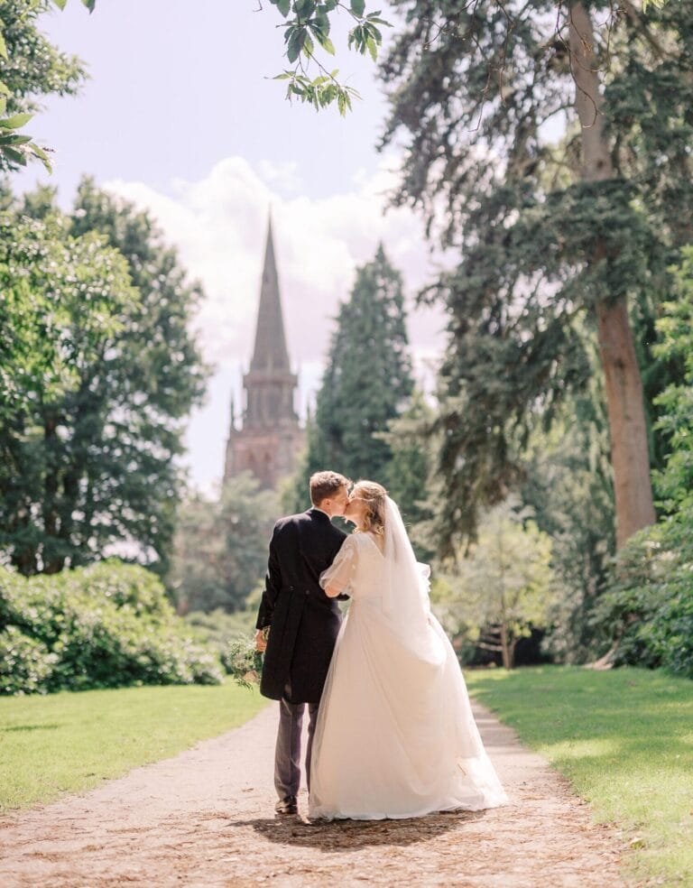 A bride and groom walk together on a path in a lush garden. The bride wears a white gown and veil, while the groom is in a dark suit. They embrace as they walk toward a tall church spire in the background. Trees and greenery surround them.