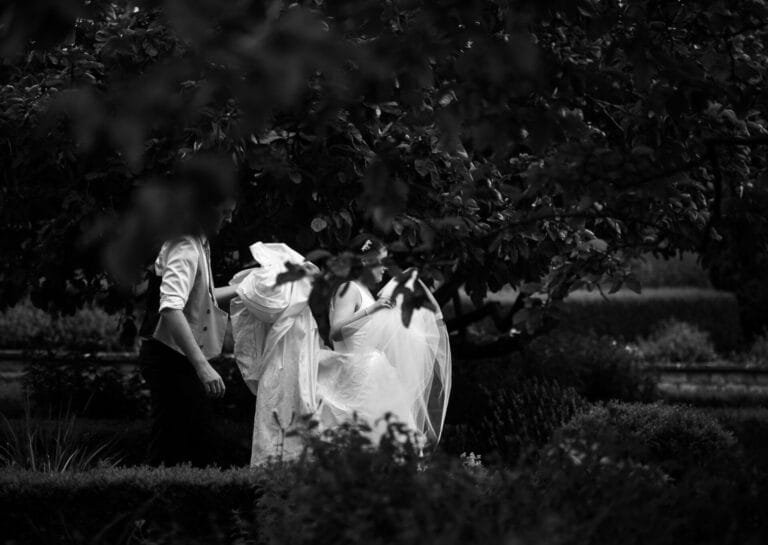 A black and white photo of a couple in wedding attire. The bride holds her dress while the groom stands nearby, both surrounded by lush foliage and greenery. The scene is intimate and serene.