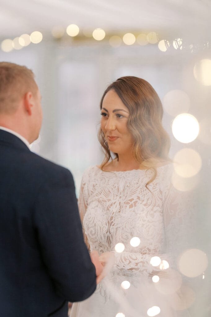 A bride and groom stand facing each other during their wedding ceremony at The Woodman Inn in Thunderbridge. The bride, in a lace wedding dress, smiles as warm, blurred lights create a soft, romantic ambiance in the foreground.