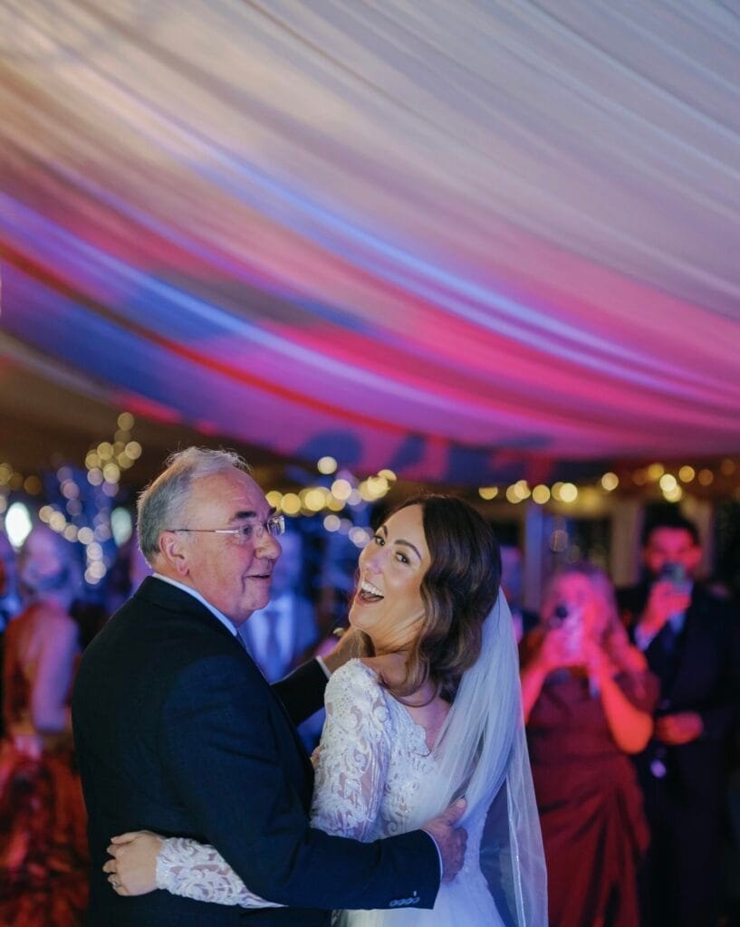 At The Woodman Inn, a bride in a white dress and veil dances joyfully with an older man in a suit. Under the ceilings colorful soft lighting, guests at this Thunderbridge celebration capture moments to cherish forever.