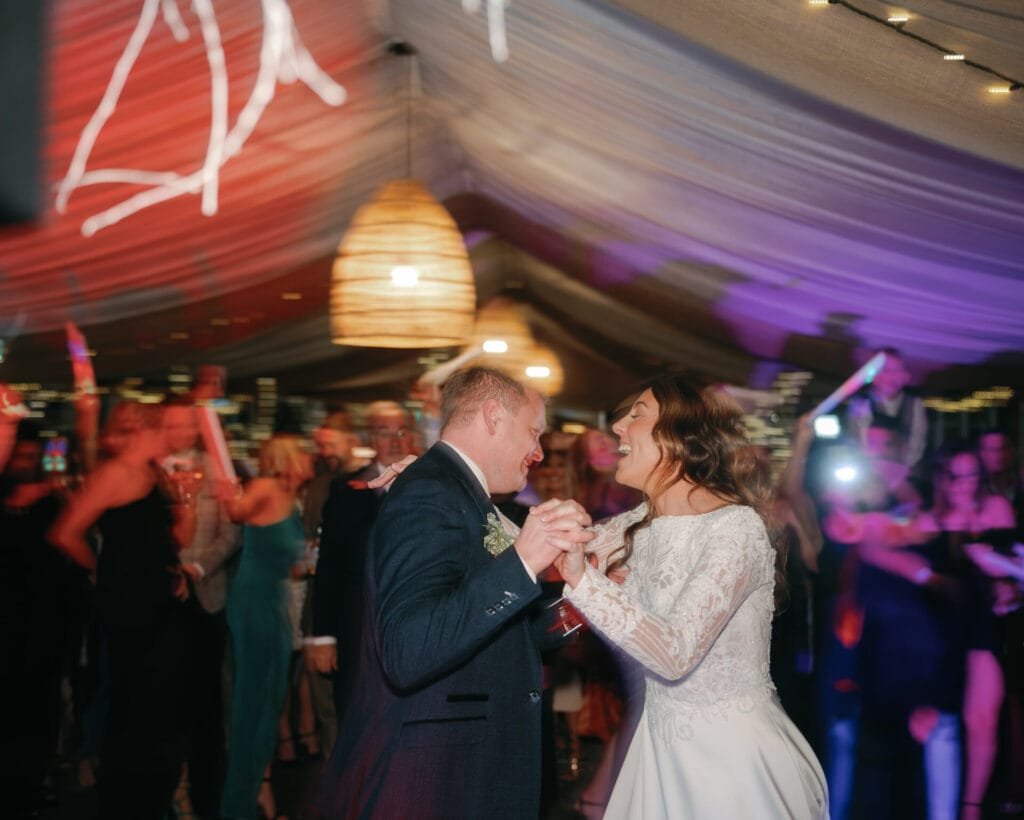 A bride and groom smiling as they dance together at their Woodman Inn wedding reception. The venue is decorated with string lights and draped ceilings. Guests in the background are holding light sticks, celebrating joyfully with their Thunderbridge wedding photographer capturing every moment.