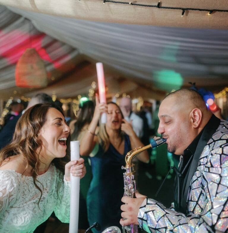 At The Woodman Inn, a bride in a white dress joyfully sings into a white plastic tube while a saxophonist in a shiny jacket plays music. Surrounded by people dancing under colorful lights, the Thunderbridge atmosphere heightens the festive celebration.