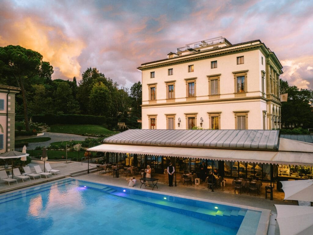 A luxurious Villa Cora with classic architectural style stands beside a serene swimming pool. Elegant outdoor seating is arranged under a canopy. The sky, painted with warm hues of a sunset, enhances the tranquil atmosphere—perfect for a destination wedding photographer to capture timeless moments.