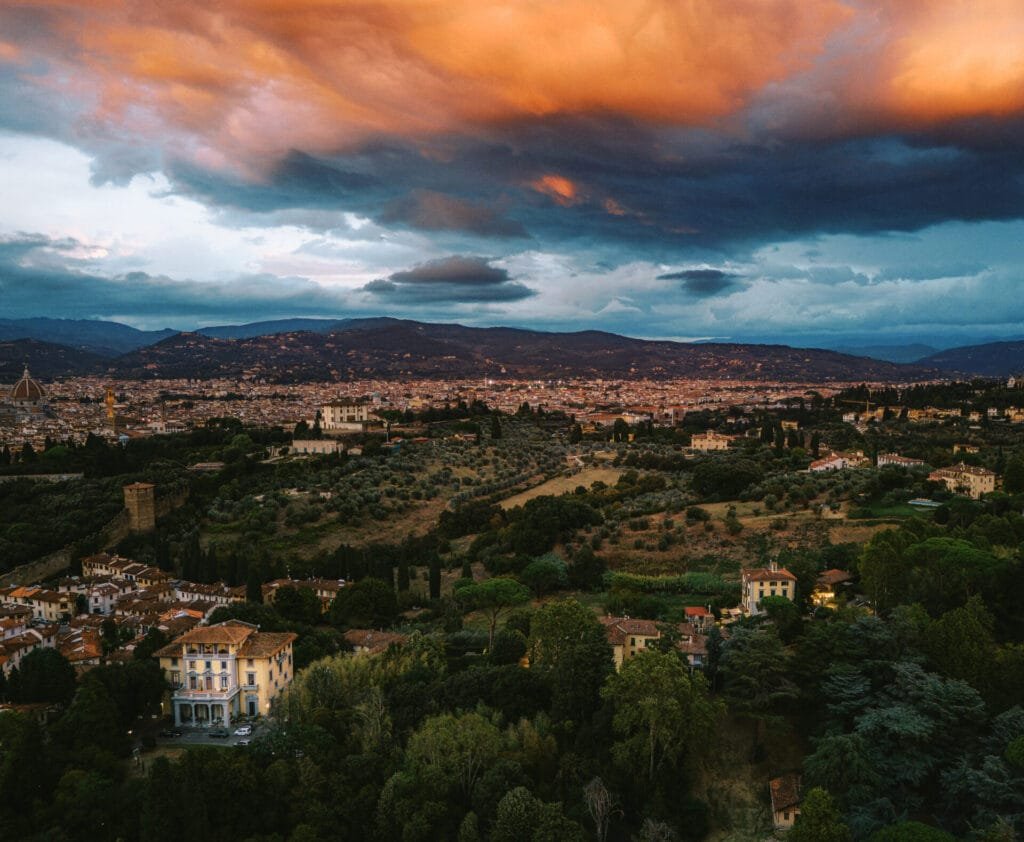 Aerial view of a Tuscan landscape with a vibrant sunset. Warm hues dance over rolling hills, scattered houses, and lush greenery. Villa Cora in Florence awaits as an idyllic destination wedding venue under this breathtaking sky for photographers capturing unforgettable moments.