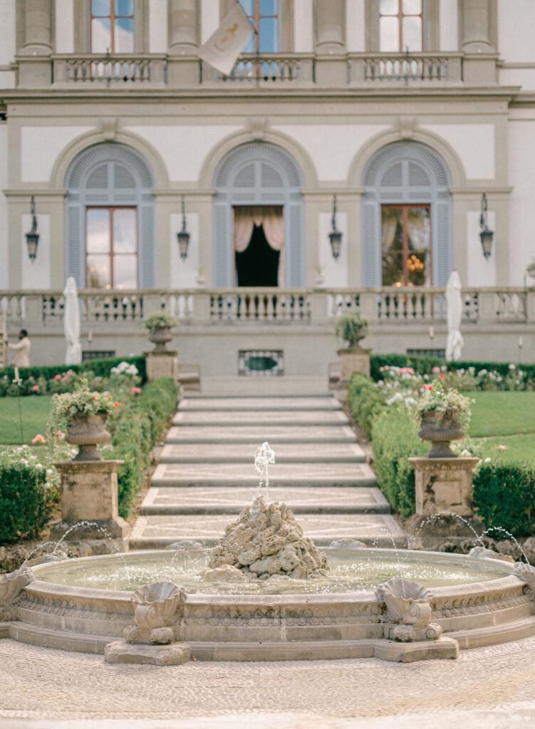 Elegant European-style garden with a central fountain graces the entrance of Villa Cora, an exquisite Florence destination. Stone steps, flanked by manicured hedges and decorative planters, lead up to the grand building—a dream setting for a wedding photographer to capture timeless moments.