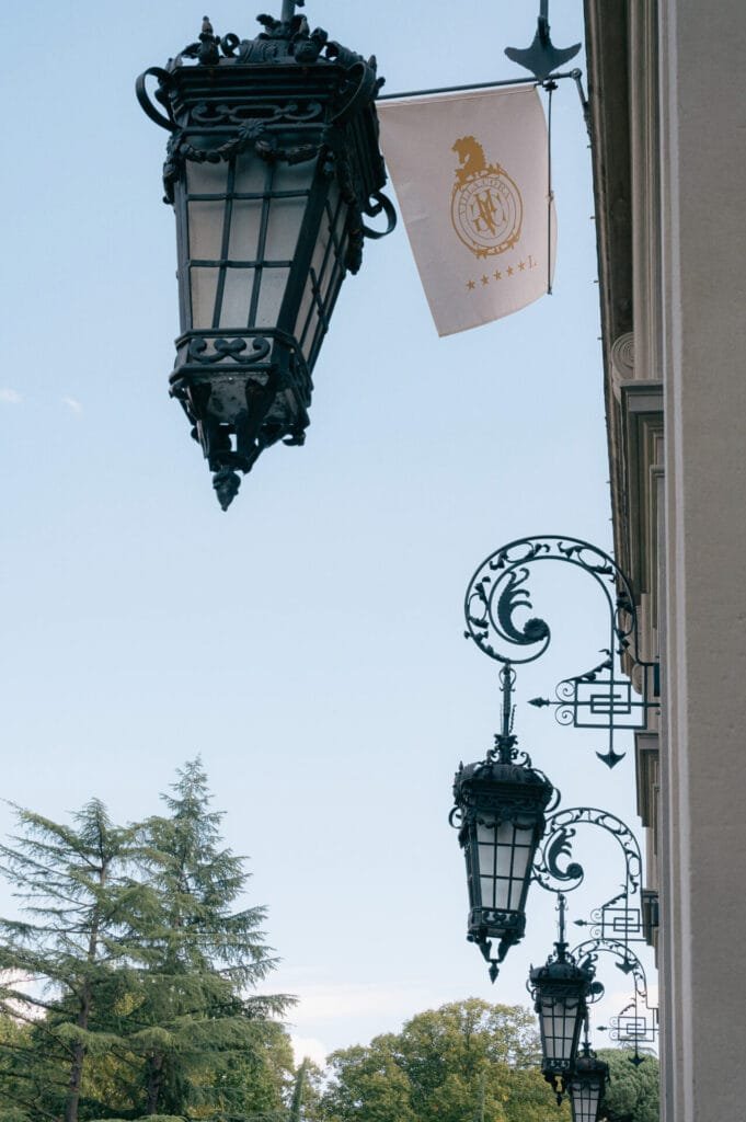 Three ornate black lanterns adorn the facade of Villa Cora in Florence, set against a vibrant blue sky. Nearby, a flag with a golden emblem and stars flutters gracefully. Green trees serve as the perfect backdrop for any destination wedding photographer capturing this enchanting scene.