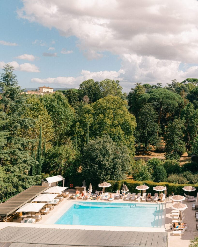 A swimming pool surrounded by loungers and umbrellas sits in a lush, forested area. In the distance, Villa Cora is visible amidst dense green trees under a partly cloudy sky—a perfect setting for a destination wedding photographer to capture unforgettable moments.