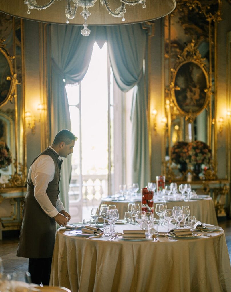 A waiter in a formal vest and apron is setting tables in an elegant restaurant at Villa Cora, where ornate decor, large mirrors, and chandeliers create a refined atmosphere. The tables are adorned with white cloths, glasses, and utensils as light streams through tall windows with green drapes.