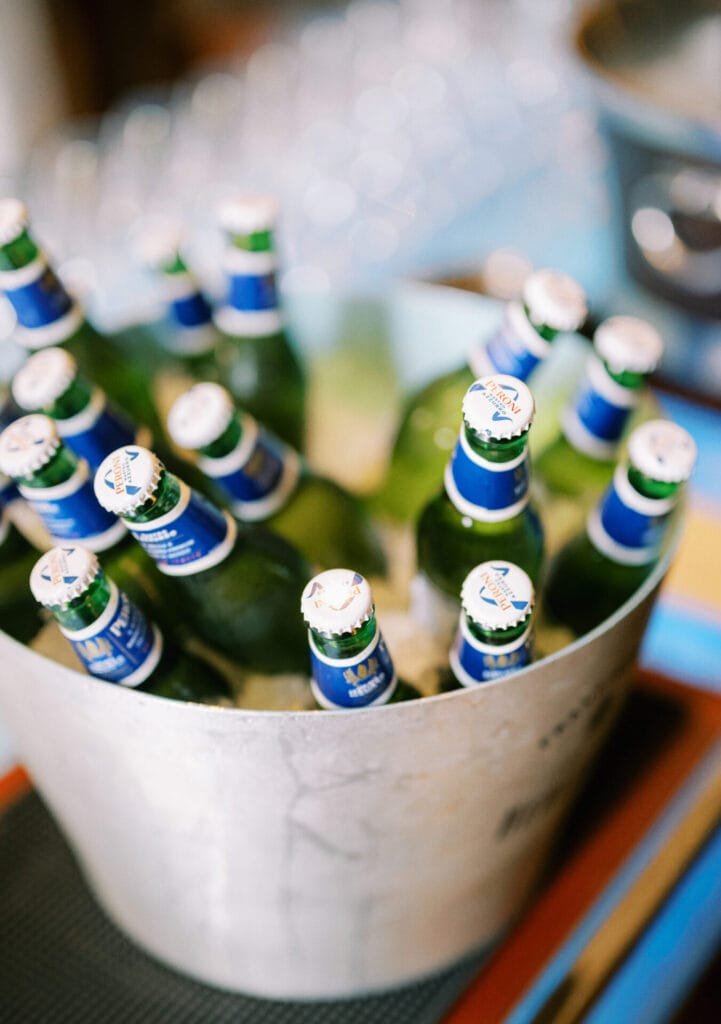 A metal bucket brimming with green glass beer bottles, each sealed with a blue and white cap, sits elegantly on a bar counter. In the background, blurred glasses whisper stories of celebrations captured by a destination wedding photographer at Villa Cora Florence.