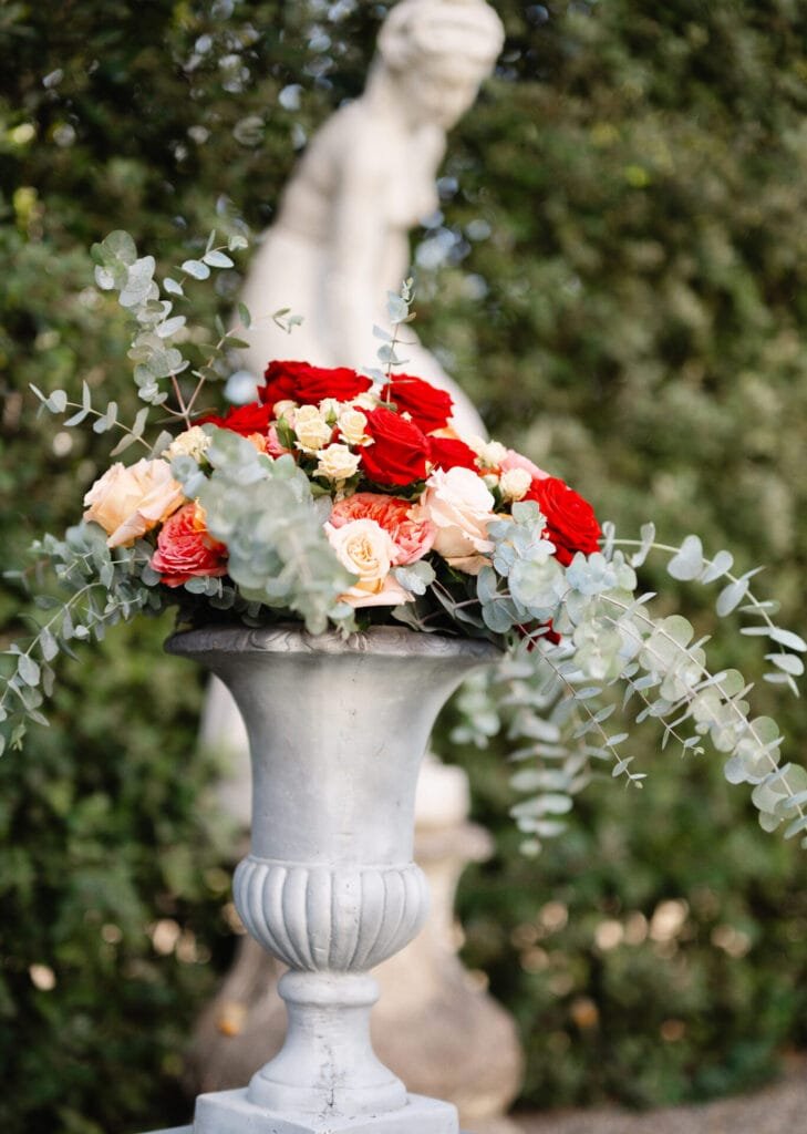 A large stone vase brimming with red and peach roses, white blooms, and greenery graces a garden setting. In the backdrop of Villa Cora Florence, a blurred statue is enveloped by lush foliage, creating an idyllic scene for a destination wedding photographer.