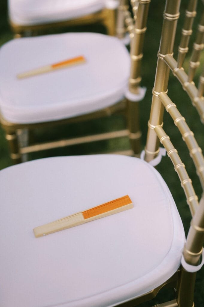 Golden chairs with white cushions are lined up in a row, each seat featuring a beige and orange folding fan, capturing the elegance of a Villa Cora Florence wedding. The grassy ground is visible in the background, creating the perfect scene for a talented photographer.