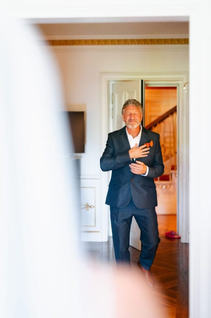 A man in a navy suit stands smiling at the doorway of Villa Cora in Florence, his hand on his chest. The background reveals part of a staircase, while the blurred foreground hints at an open door or curtain—a perfect snapshot by a wedding photographer.