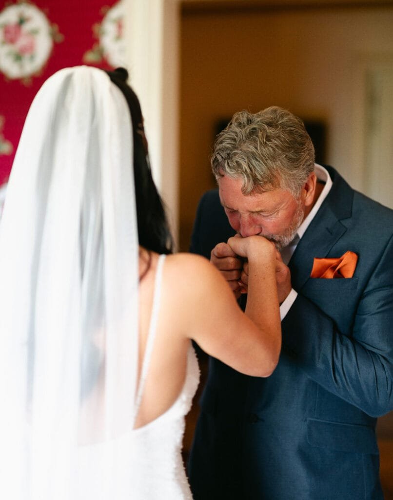 A bride in a white dress and veil has her hand kissed by a man with gray hair, dressed in a blue suit and orange pocket square. Captured by a Villa Cora Florence wedding photographer, the moment is set against a floral-patterned wall.