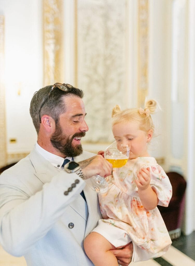 In the elegant ambiance of Villa Cora, a bearded man in a light suit joyfully holds a glass of orange juice for a young girl with blonde pigtails and a floral dress. Both are smiling, captured beautifully by the wedding photographer amidst the ornate decor.