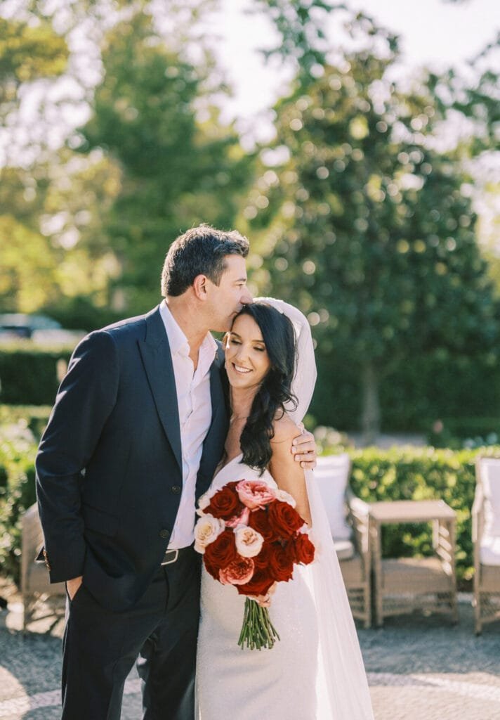 Bride holding a bouquet of red and pink roses, smiling, while a man in a suit gently kisses her head. They are outdoors at Villa Cora Florence, surrounded by greenery and dappled sunlight—a perfect moment captured by the wedding photographer.