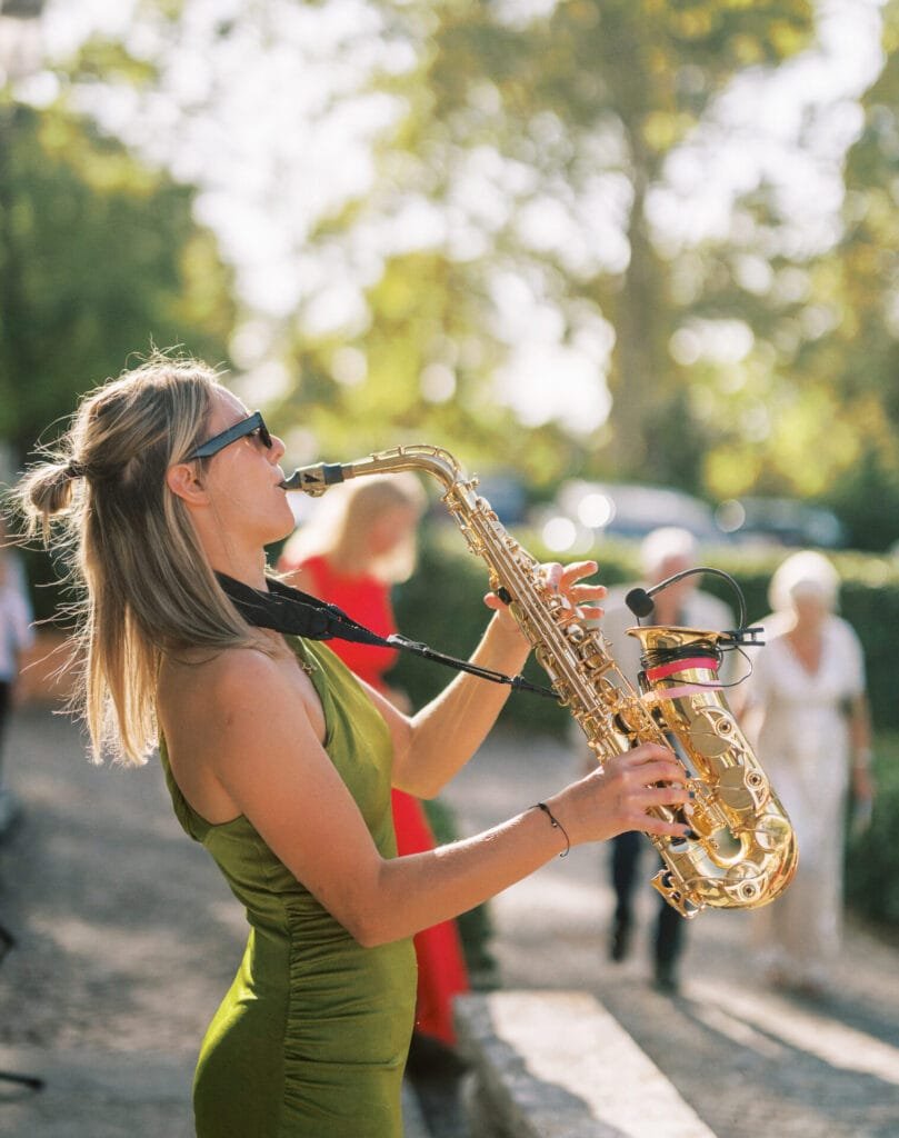 A woman in a green dress and sunglasses plays a saxophone outdoors, captured beautifully by a Villa Cora Florence wedding photographer. The sunny scene with blurry trees and people in the background adds to the vibrant, lively atmosphere.