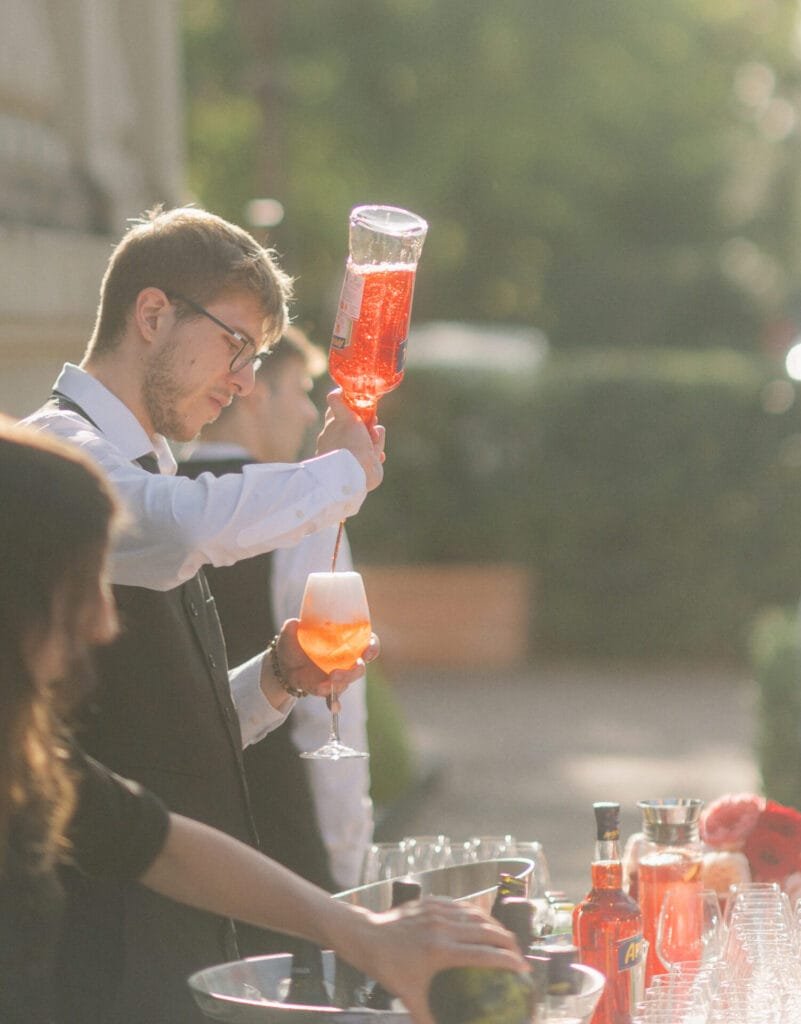 At an outdoor bar, a bartender at Villa Cora pours a pink drink from a bottle into a glass. Sunlight dances through the trees, casting warmth and adding charm to the scene, reminiscent of moments captured by a Florence wedding photographer. Assorted bottles and glasses adorn the bar.