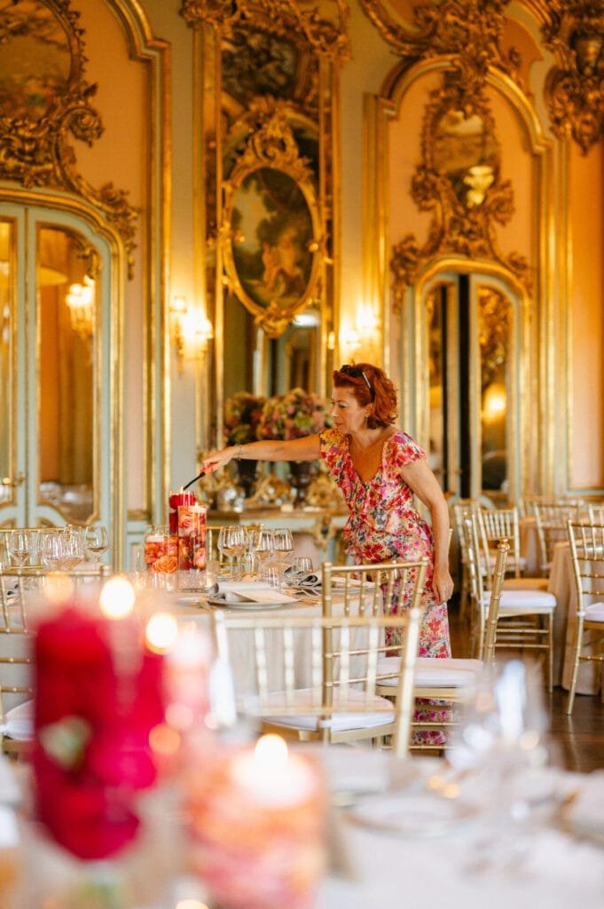 In a lavishly decorated room at Villa Cora, Florence, a woman in a floral dress elegantly arranges red candles on a table set for an event. The ornate mirrors and gold accents create the perfect backdrop for any wedding photographer.