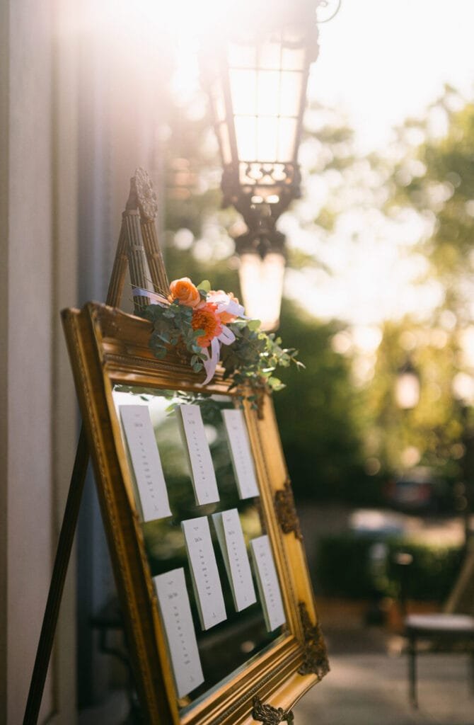 An ornate frame on an easel holds a seating chart with several paper cards. Sunlight filters through a lantern above, casting a warm glow. A small floral arrangement decorates the top left corner of the frame. Captured by a Villa Cora Florence wedding photographer, trees are blurred in the background.