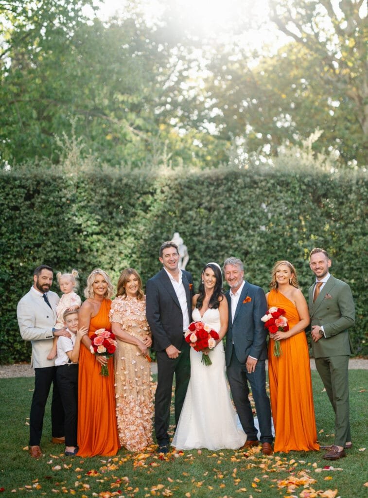 A wedding party poses outdoors, captured by a skilled Villa Cora Florence wedding photographer. The bride in a white gown and groom in a black suit stand at the center, surrounded by family members. Bridesmaids don orange dresses, while a young child is held by a man on the left. Trees and a statue add to the serene backdrop.