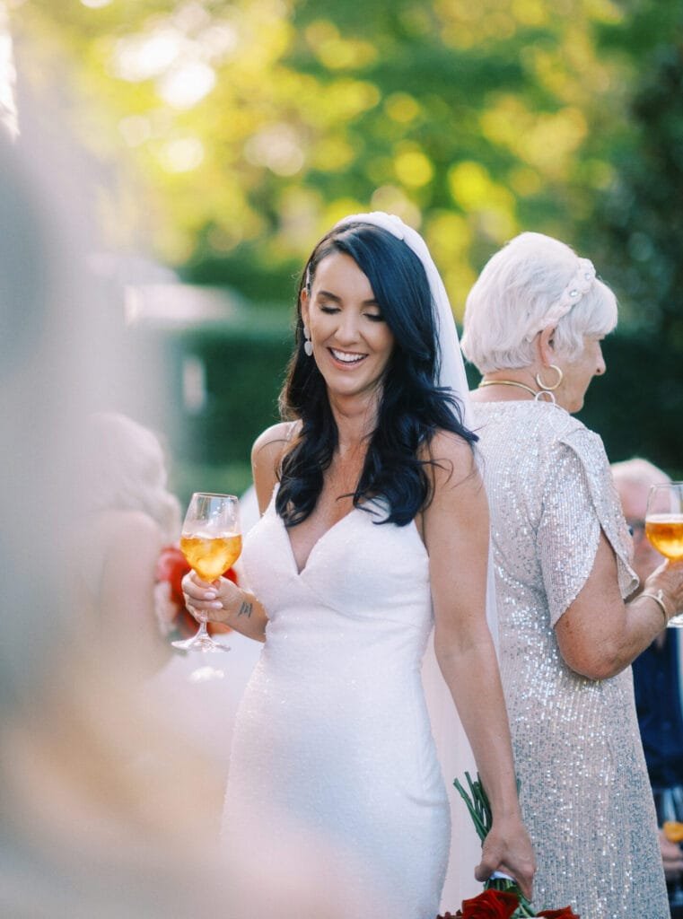 A bride in a white dress and veil smiles, holding a drink in a wine glass at Villa Cora Florence. She is outdoors, surrounded by guests with a blurred background of greenery. Beside her, another woman in a silver outfit also holds a drink, perfectly captured by the wedding photographer.