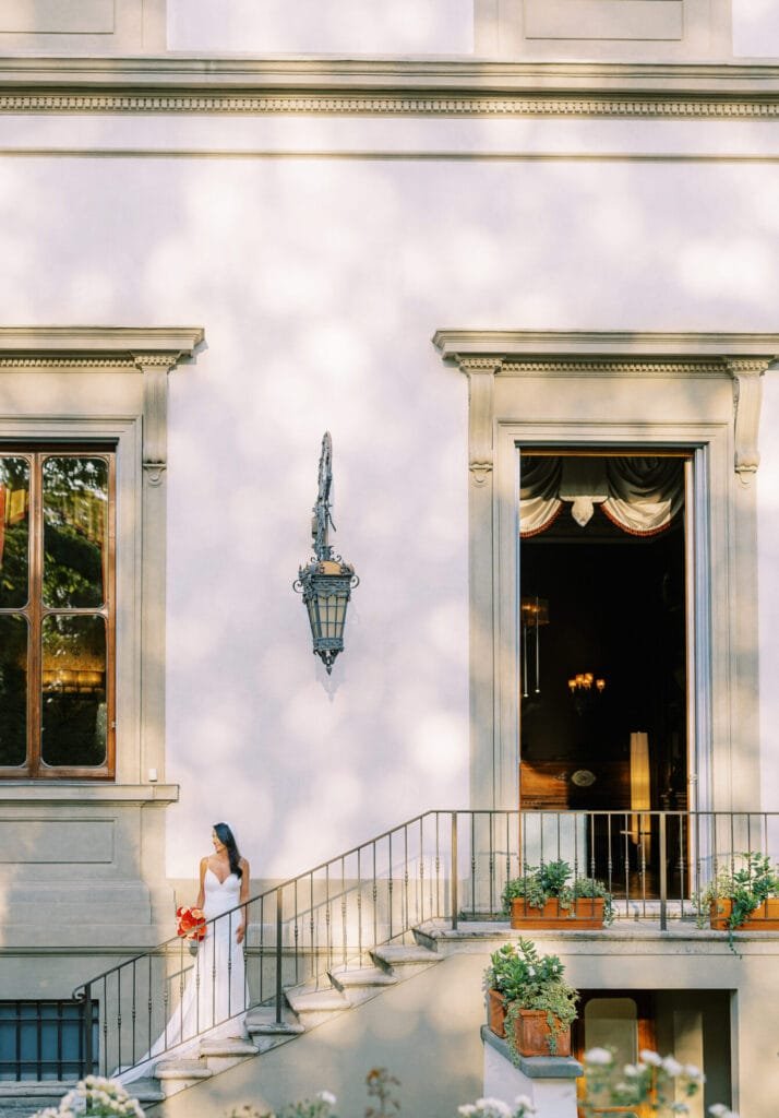 A bride in a white dress stands on an outdoor staircase at the majestic Villa Cora, adorned with classical architectural details. She holds a bouquet and gazes thoughtfully. Wrought iron railings and potted plants line the stairs, perfect for any Florence wedding photographer to capture.