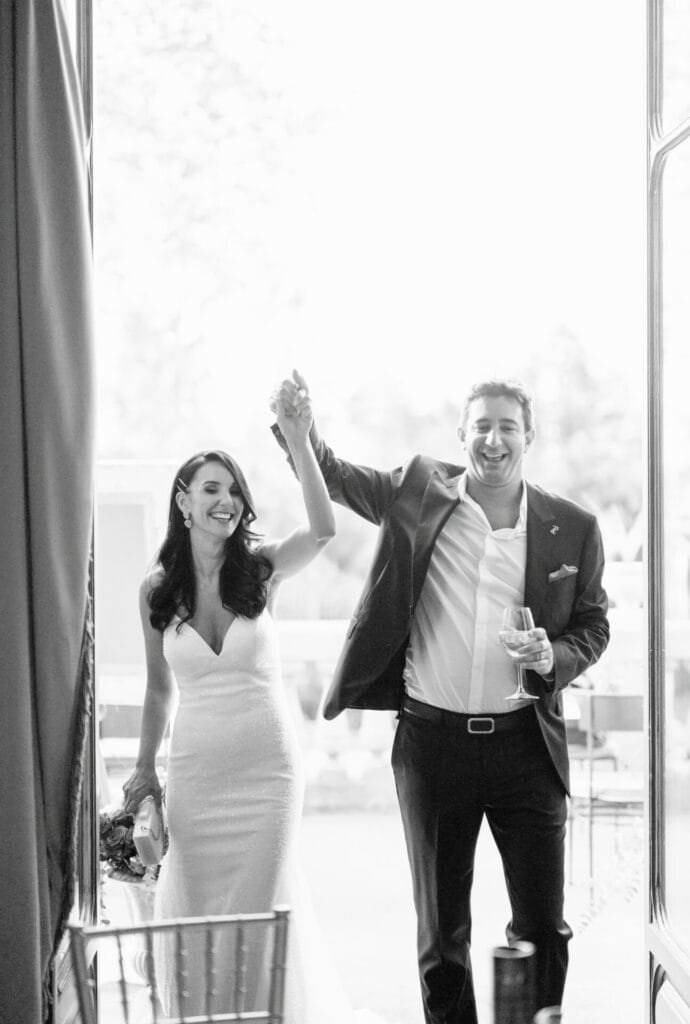 A bride and groom walk hand in hand, joyfully celebrating at Villa Cora. The bride wears a fitted, strapless gown, while the groom is in a suit, holding a drink. Captured by a Florence wedding photographer, they exude happiness as they enter the venue. Black and white photo.