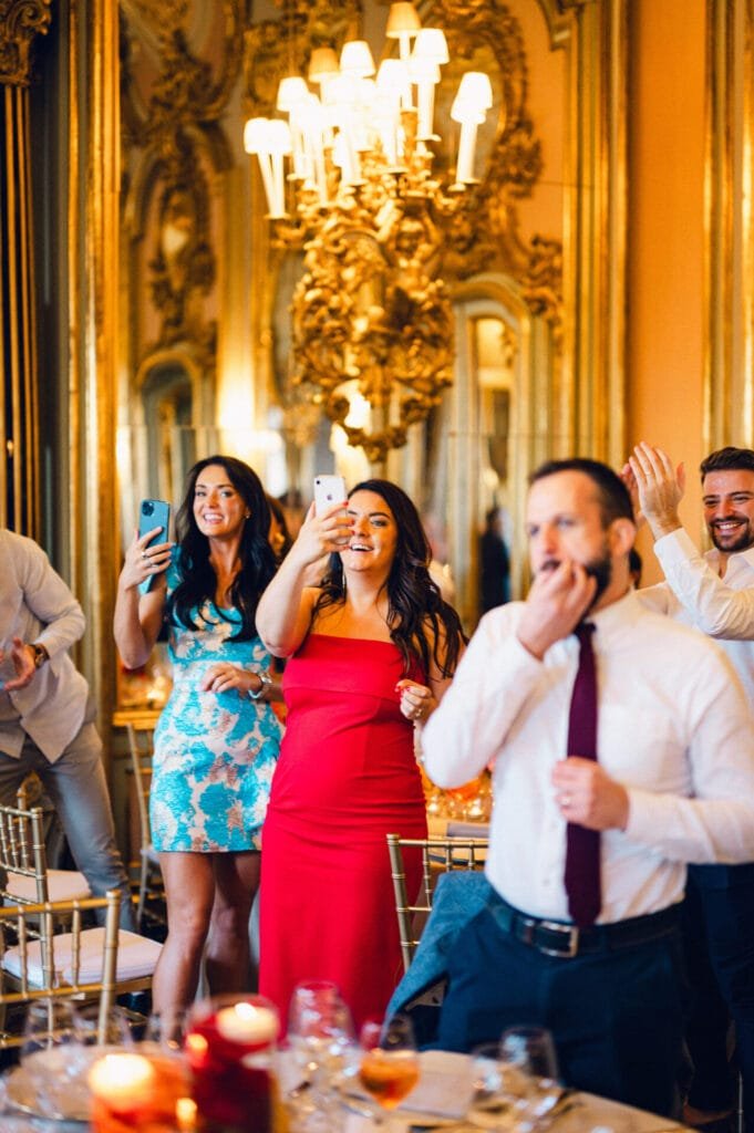 At a lavish Villa Cora Florence event, the joyous group claps and captures memories amid golden decor and a grand chandelier, embodying the celebratory spirit that delights any wedding photographer.