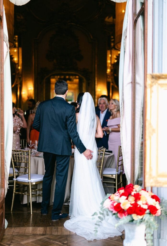 In a luxurious room at Villa Cora, a bride and groom stand gracefully, seen from behind. The bride wears a long white gown and veil, the groom in a dark suit. Guests enjoy elegant decor at round tables while red and white flowers bloom in the foreground, captured by a Florence wedding photographer.