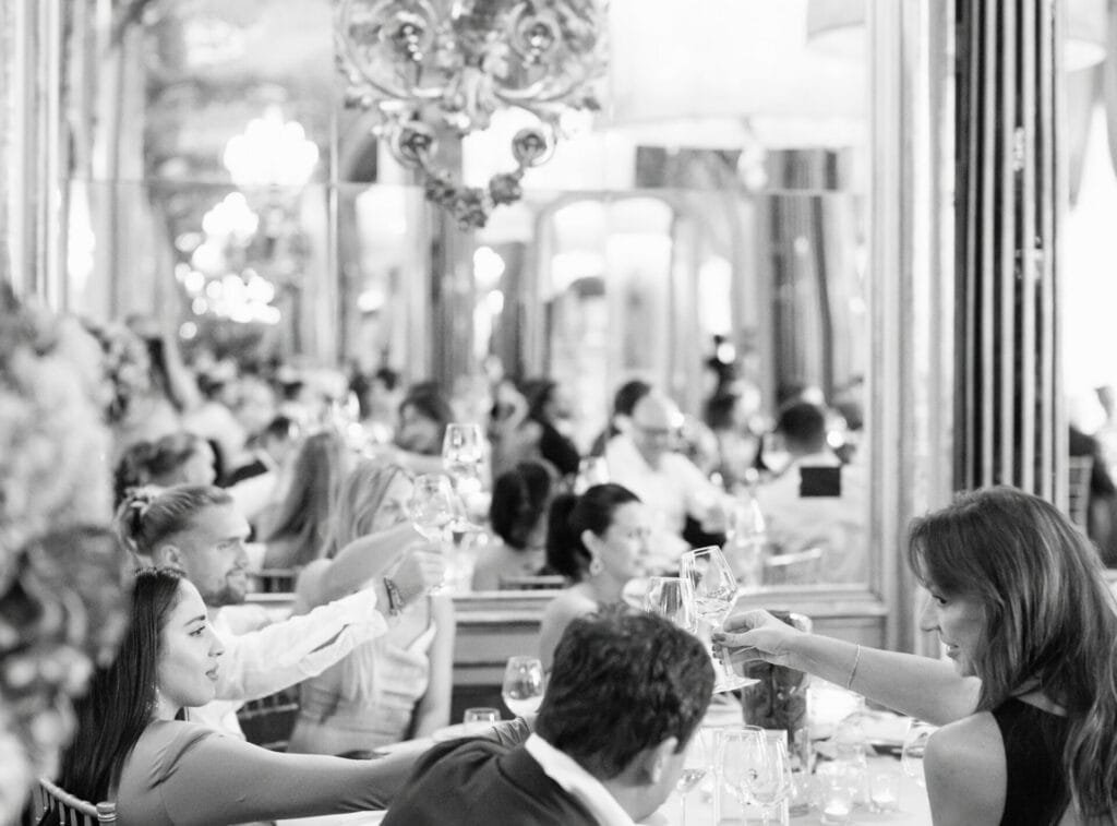 Black and white photo of a lively dining scene in an ornate room with mirrors and chandeliers at Villa Cora in Florence. Guests are seated at tables, raising glasses in a toast, captured perfectly by the wedding photographer to create a festive and elegant atmosphere.