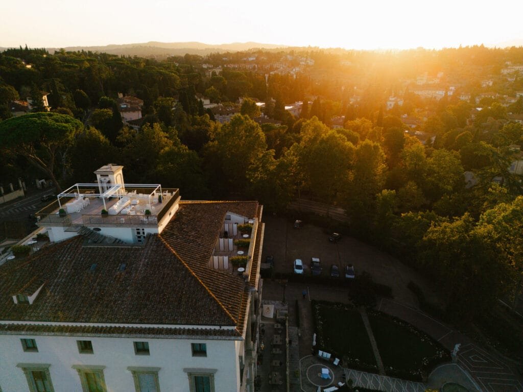 Aerial view of the magnificent Villa Cora at sunset, perfect for a Florence wedding. The rooftop terrace offers breathtaking panoramas, with lush green trees and hilly landscapes stretching out beneath the warm, golden glow of the low sun. An idyllic backdrop for any photographers lens.