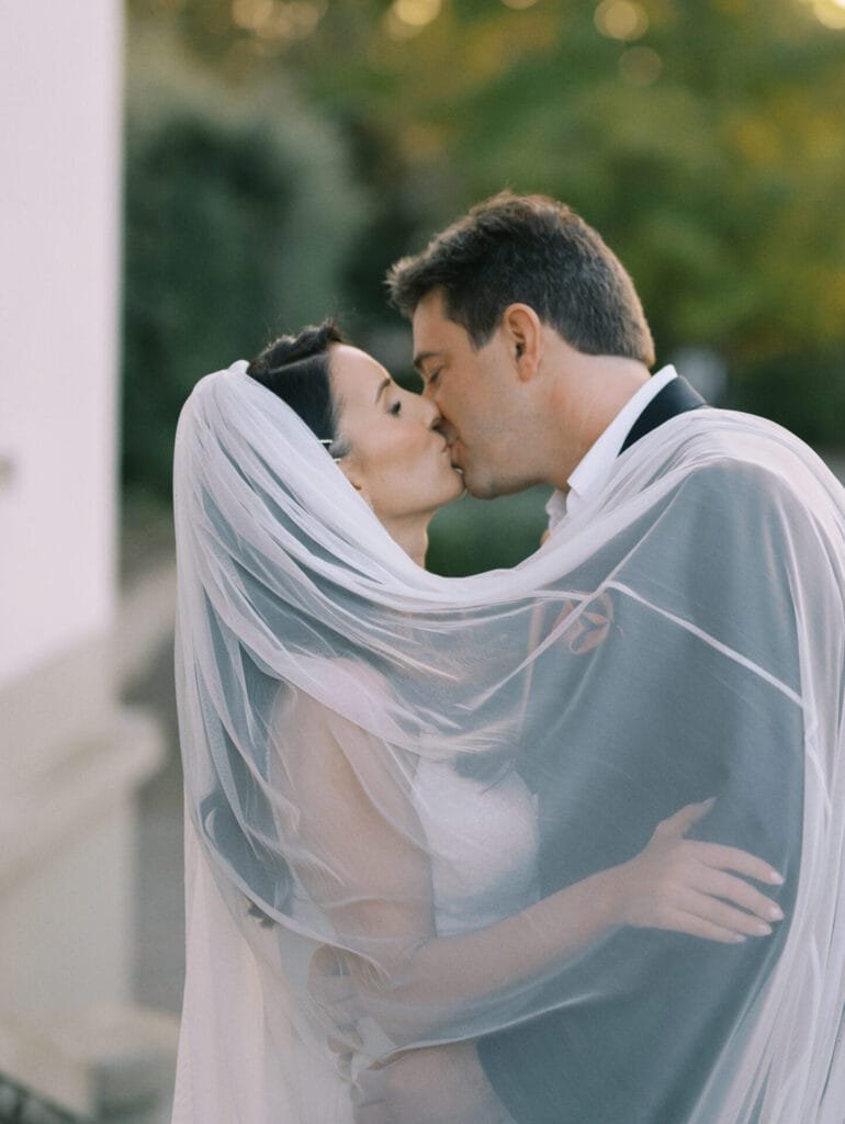 A bride and groom share a kiss under a sheer veil at Villa Cora. The bride is in a white dress, and the groom is in a suit. With greenery softly blurred in the background, the Florence wedding photographer captures this romantic atmosphere beautifully.