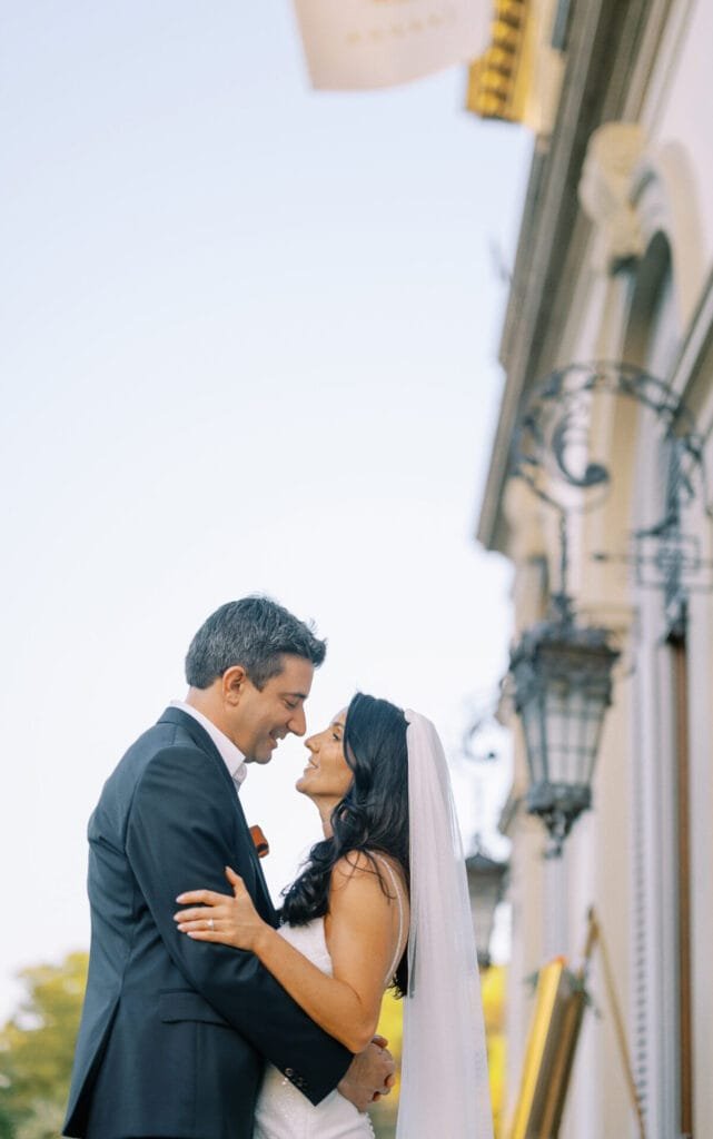 A bride and groom stand closely, smiling at each other. The bride wears a white dress and veil, while the groom is in a black suit. Captured by a Villa Cora Florence wedding photographer, they pose near ornate lanterns under a clear blue sky.