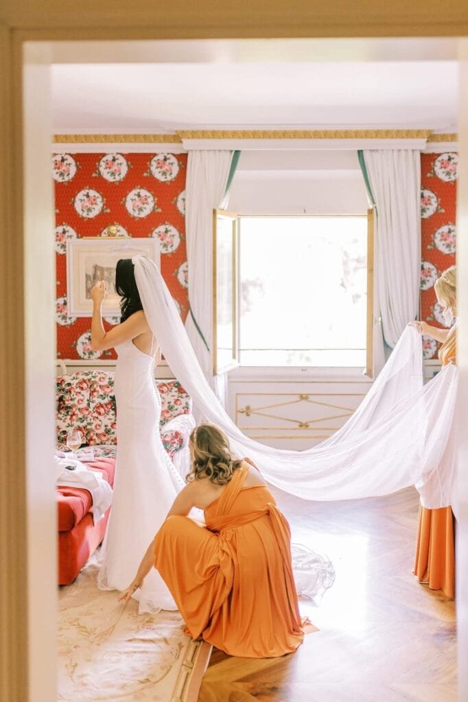 A bride in a white wedding dress stands in a room with floral wallpaper. Her veil is held by two bridesmaids wearing orange dresses. The large window in the background lets in natural light, illuminating the scene—a perfect moment captured by a Florence destination wedding photographer.