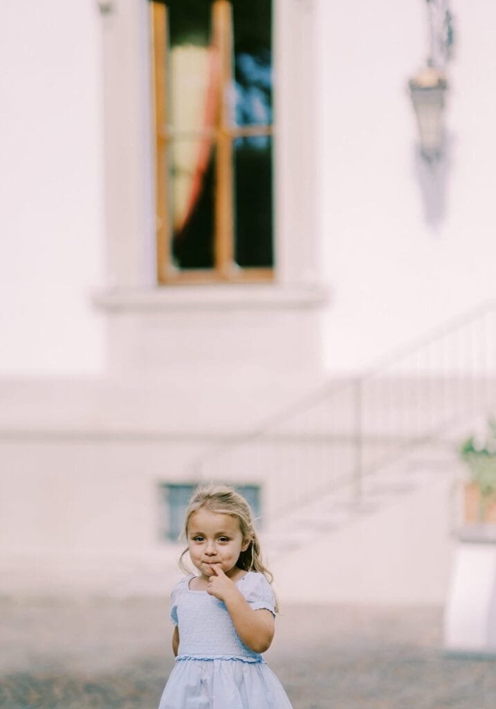 A young girl in a light blue dress stands outdoors, touching her chin thoughtfully. She is poised in front of a white building with a window and staircase railing in the background, evoking the elegance captured by a Florence destination wedding photographer.