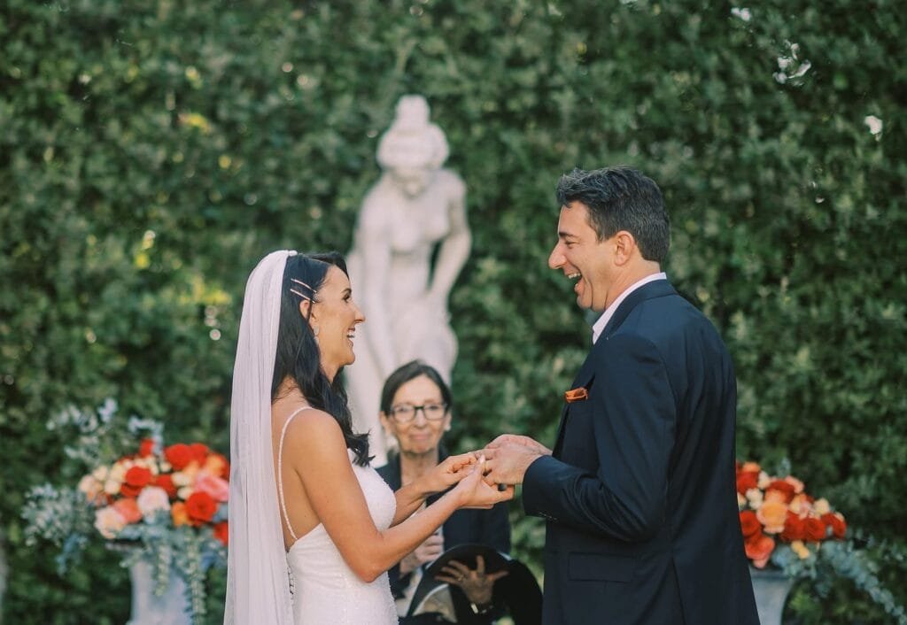 At a stunning outdoor ceremony, captured by a Florence destination wedding photographer, the bride and groom exchange rings. Smiling at each other before an officiant, they stand amidst a lush green hedge, a white statue, and vibrant red and orange floral arrangements.