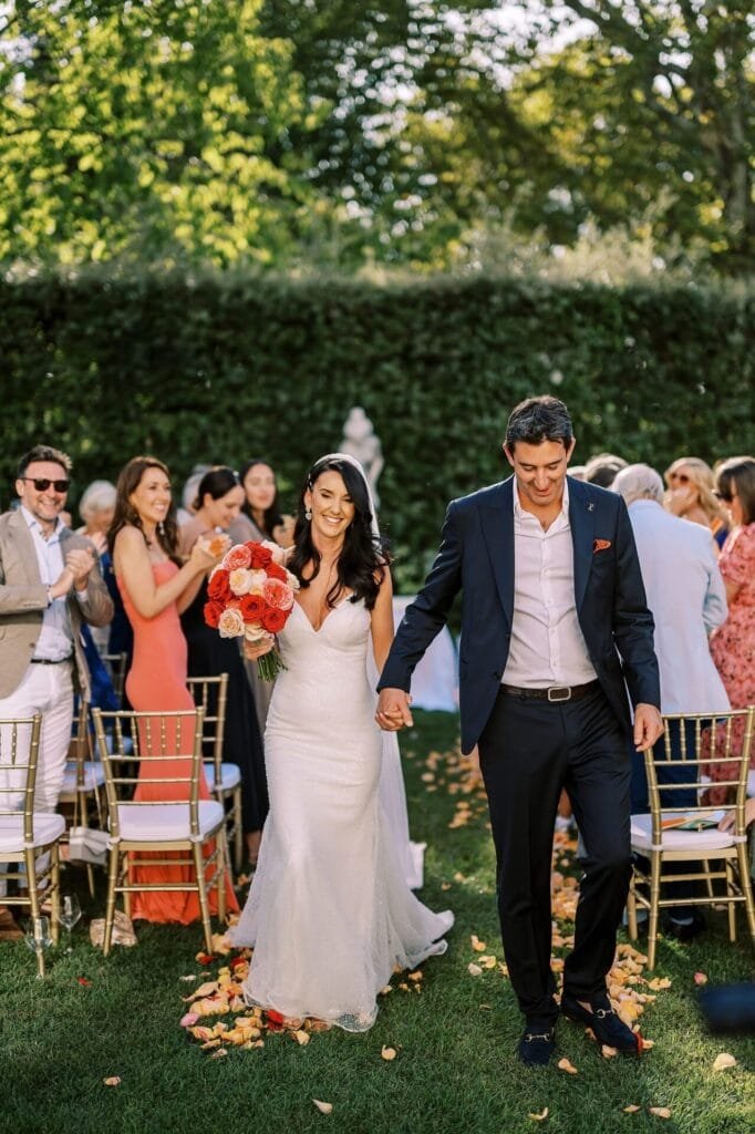 A bride and groom smile as they walk down an outdoor aisle, surrounded by guests clapping and cheering. The bride holds a bouquet of orange and pink flowers. With sunlight filtering through lush greenery, its a perfect scene for a Florence destination wedding photographer to capture their joy.